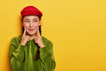 Pretty young Asian model keeps both fore fingers on cheeks, looks with dreamy expression aside, has minimal makeup, wears red beret, green velvet hoody, poses over yellow wall with blank space