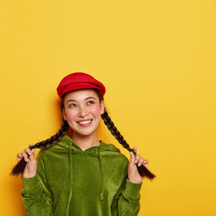 Fascinated tender young Korean woman shows long braids, has dreamy pensive expression, dressed in stylish vivid clothes, poses over yellow studio wall, blank space above, stays happy and pleased