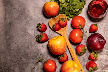 Fresh organic red themed fruit, vegetables, wine, and wooden heart shaped spoon, laying on concrete background countertop. Captured from above. 