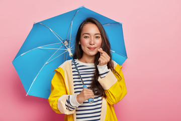 Weather, autumn and people concept. Friendly looking brunette lady make like hand sign, shapes little heart near cheek, has Asian appearance, dressed warmly during wet rainy day, holds umbrella