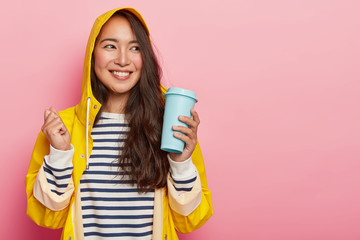 Positive woman with long dark straight hair, raises clenched fist, holds takeaway coffee, dressed in striped jumper, yellow raincoat, enjoys hot drink during cold rainy autumn day, feels calm, relaxed