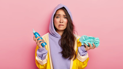 Image of frustrated young woman bites lower lip, holds spray and handkerchief, looks nervously at camera, feels sick, caught cold during rainy day, wears purple hoody. Autumn, people, health problem