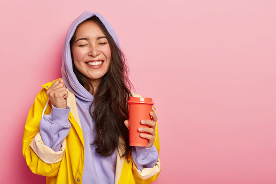 Overjoyed pleased Asian woman raises clenched fist, keeps eyes closed, drinks hot beverage from flask, rejoices happy moment, wears warm waterproof clothes, ready for walk in forest or park. - Powered by Adobe