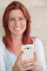 Charming smiling redhead woman sitting against an old wall in the process of sending messages by mobile phone