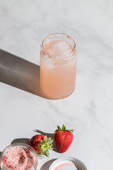 A strawberry lemonade drink in a can like glass standing on a marble countertop. Direct sunlight from the aide casting a hard shadow on the countertop. Bottom is whole strawberry with strawberry sugar - 289754638