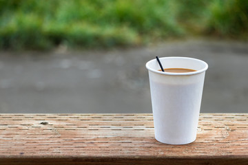 A disposable paper cup of coffee placer on a wooden table over a nature background, outdoors, close up.