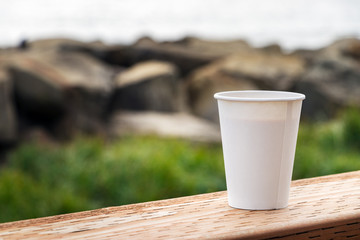 A disposable paper cup of coffee placer on a wooden table over a nature background, outdoors, close up.