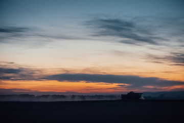 Silhouette of harvester machine to harvest wheat on sunset. Combine harvester driving on field on sunrise. Beautiful dawn sky above wheat field. Combine working in dusk. Wonderful twilight landscape.
