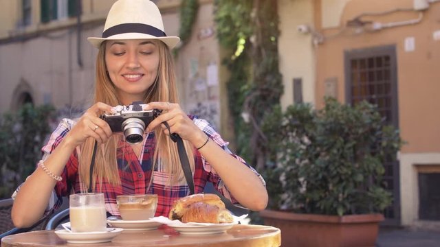 Young blonde woman with blue eyes having breakfast in typical Italian bar outside in historical neighborhood Trastevere in Rome, Italy. Taking pictures with vintage camera. Cappuccino, coffe and