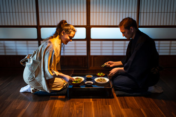 Young couple seiza sitting on floor pillows with Japanese ryokan kaiseki dishes, eating soba noodles, natto, edamame and soy sauce on table by sliding paper doors - obrazy, fototapety, plakaty