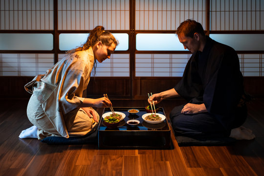 Young couple seiza sitting on floor pillows with Japanese ryokan kaiseki dishes, eating soba noodles, natto, edamame and soy sauce on table by shoji sliding paper doors