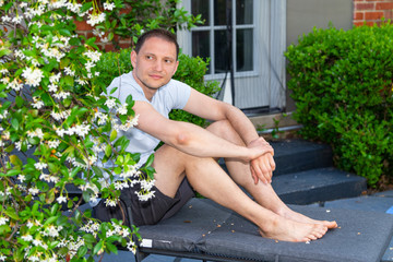 Young man sitting on patio lounge chair in outdoor spring flower garden in backyard of home plants