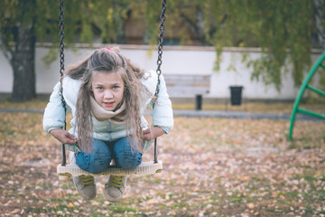 Cute girl dressed in down jacket sitting on swing on the playground on cool autumn day.