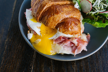 Croque Madam croissant breakfast sandwich with fried egg with runny yolk, pork ham, and melted cheese served on ceramic plate with side salad. Rustic wood table surface.