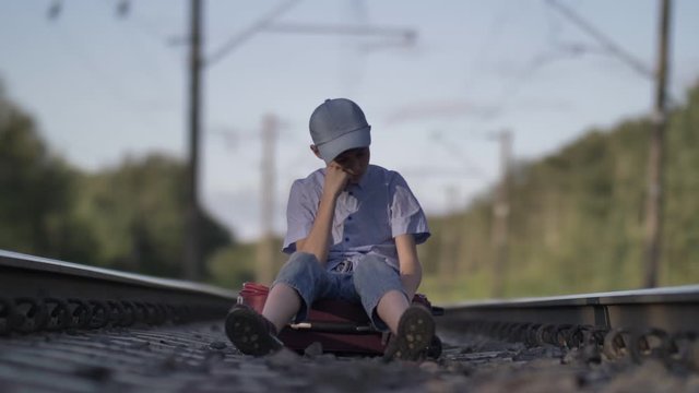 sad boy in a cap sits on a suitcase on the railway outdoors