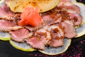 Edged pieces with caramelized pear on a black ceramic plate. Baked meat sliced with thin pieces