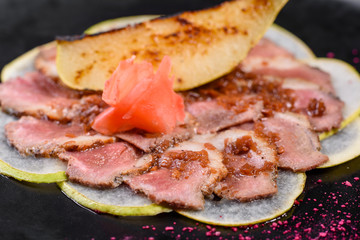 Edged pieces with caramelized pear on a black ceramic plate. Baked meat sliced with thin pieces