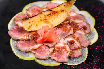 Edged pieces with caramelized pear on a black ceramic plate. Baked meat sliced with thin pieces