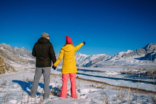 Cute young couple in bright down jackets on the backdrop of beautiful winter mountain scenery with copy space. Christmas holidays in mountains. Man and woman looking at snowy peaks.