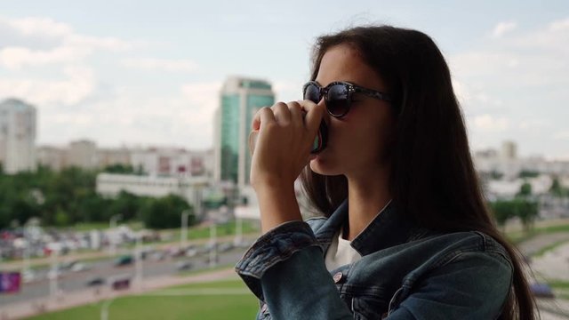 teenager with dark long hair in denim jacket and sunglasses drinks coffee looking at city from balcony close slow motion