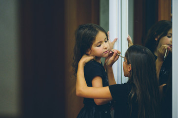 older sister making dark makeup with ink pencil on her little caucasian girl sister face preparing to Halloween holiday party