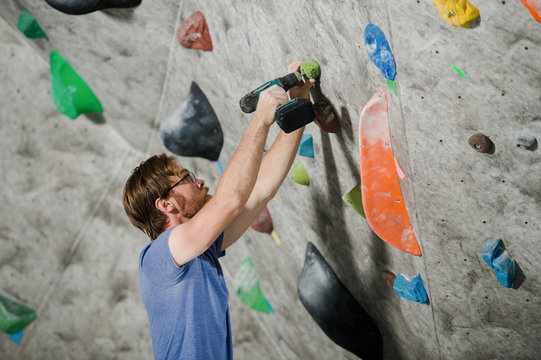 active young bearded man in glasses building climbing wall with color holds indoors using drill tool