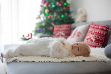 Curious little toddler boy, dressed in handknitted white overall, lying on the couch playing with gifts