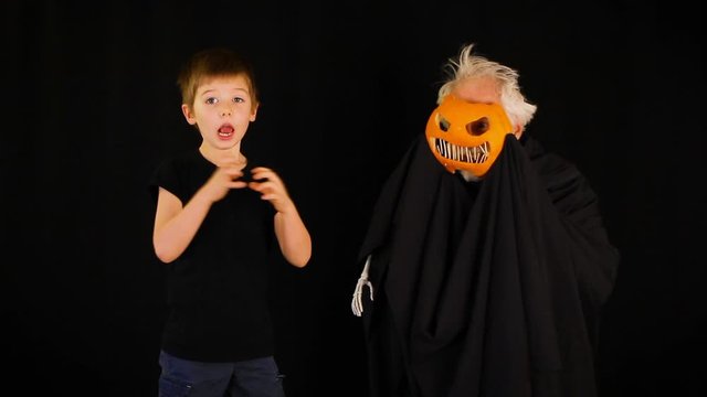 An overdramatic boy pretends to be scared  by waving hands in the air with wide eyes and an open mouth, of his grandfather who is dressed in a pumpkin head costume for Halloween. Black background.
