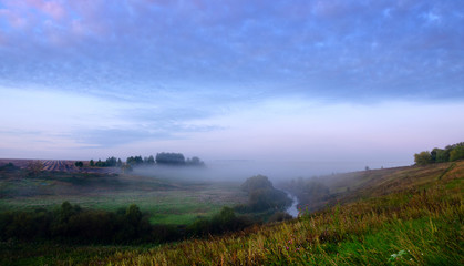 Tranquil hazy landscape with small river and hills covered by morning fog