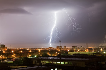 Naklejka na ściany i meble A powerful lightning strike hits the city at night. A strong lightning strike over a dark gray sky hits the ground, illuminating the industrial area around.