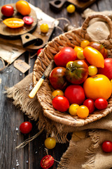 Fototapeta na wymiar homegrown assorted red, yellow, orange tomatoes in wicker straw basket stands on sackcloth on rustic wooden table, autumn harvesting, selective focus