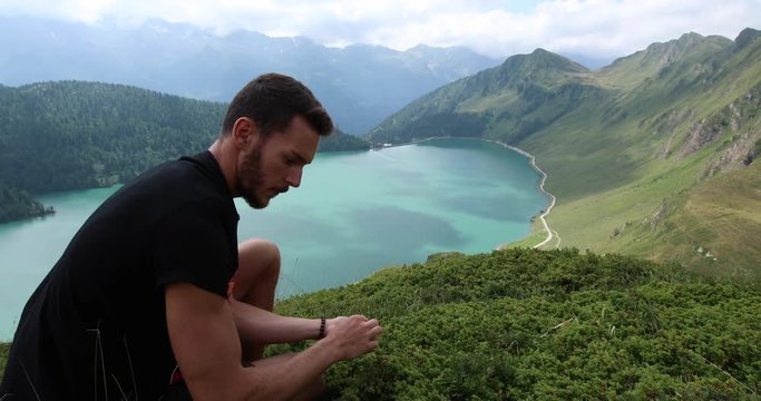 Young man harvesting blueberries during a hike in the mountains, behind him Lake Ritom.