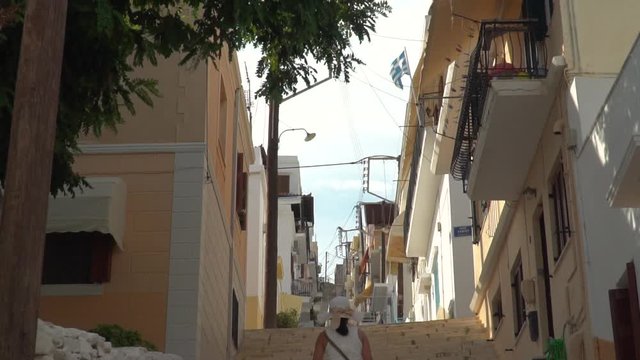 Tilt up from girl in summer dress to Greek flag walking up steps in narrow alley in Syros SLOW MOTION