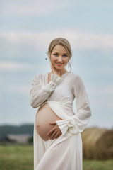Fototapeta na wymiar Pregnant girl walks in a field near haystacks in a long white dress, a woman smiles and holds her hands over her stomach. Girl is expecting the birth of a baby in the ninth month of pregnancy