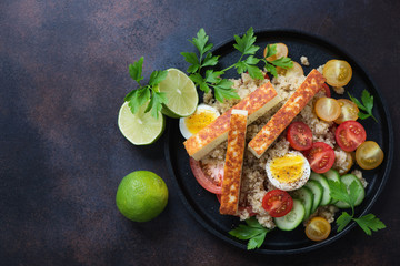 Metal tray with quinoa, roasted cheese, tomatoes, cucumber and eggs. Above view on a dark brown metal surface with space