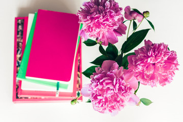 bouquet of pink peonies, a stack of books, pencils and a notebook on a white table, top view.