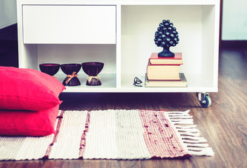 Cozy home interior decor, burning spa aroma candles in coconut shell, metal vase, pile of books, on white glass nightstand near a multi-colored rug with decorative pillows background.