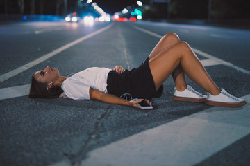 Woman in earbuds lying on night road listening to music in earphones