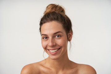 Indoor portrait of joyful young female smiling sincerely to camera, looking beautiful without make-up, standing over white background