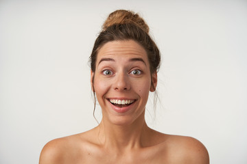 Indoor close-up of surprised young pretty woman with casual hairstyle standing over white background, looking to camera with raised eyebrows and amazed face