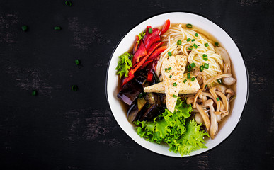 Asian vegan noodles soup with tofu cheese, shimeji mushrooms and fried eggplant in bowl on dark background. Top view