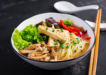 Asian vegan noodles soup with tofu cheese, shimeji mushrooms and fried eggplant in bowl on dark background.