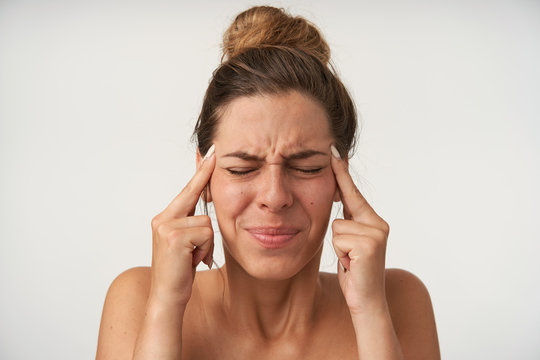Close-up of young pretty woman with painful face, posing over white background with index fingers at temples, closing eyes because of headache