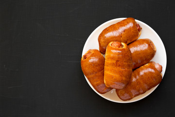 Homemade beef sausage kolache on a white plate on a black surface, top view. Flat lay, overhead, from above. Copy space.