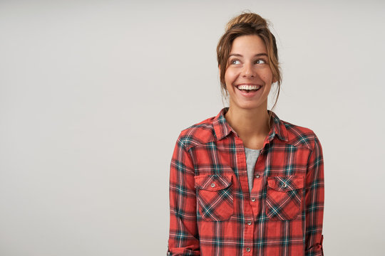 Happy young woman with casual hairstyle posing over white background in checkered shirt, looking aside cheerfully with broad sincere smile