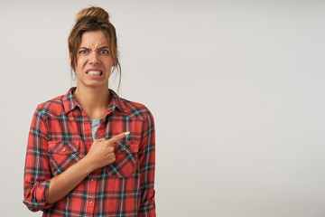 Indoor shot of young female showing with forefinger aside, wearing checkered shirt and natural make-up, pouting and frowning, showing disgust on her face