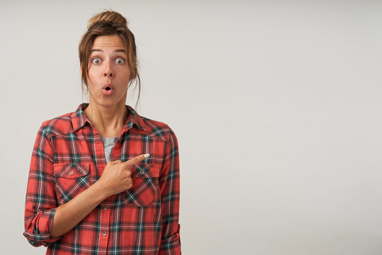 Studio photo of young amazed female in casual clothes posing over white background, pointing with index finger aside, looking to camera with eyes wide opened