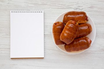Homemade beef sausage kolache on a white plate, blank sheet of paper on a white wooden background, top view. Copy space.