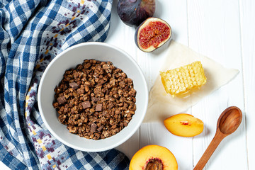 Chocolate granola in a white bowl in a composition with honeycombs, a spoon, figs and peach. Healthy breakfast food. Making breakfast. Gluten free