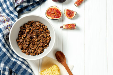 Chocolate granola in a white bowl in a composition with honeycombs, a spoon, figs on white wooden background. Healthy breakfast food. Making breakfast. Gluten free